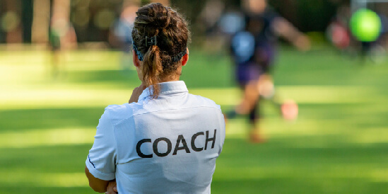 Person looking a grass wearing a white t-shirt with coach written on the back.