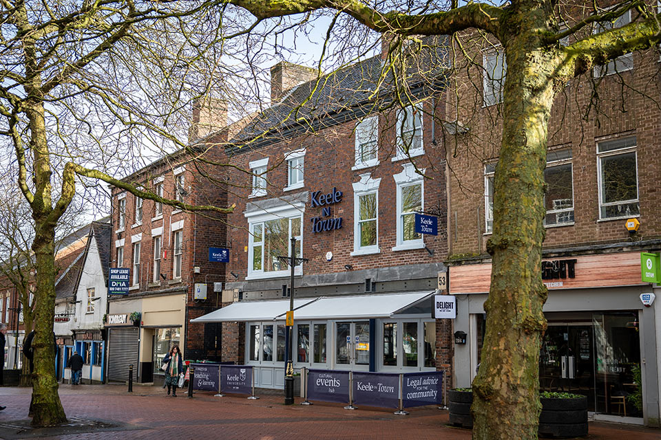 A row of brick buildings housing shops and businesses, including Keele in Town, along a pedestrian street lined with leafless trees.